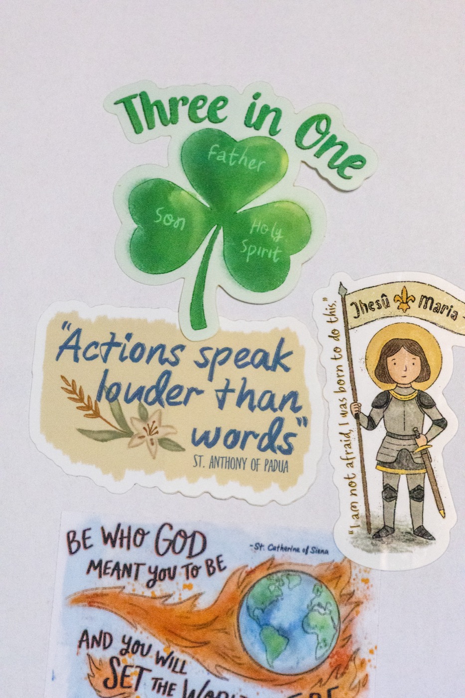 Original saint stickers — a Trinity shamrock, St. Anthony's 'actions speak louder than words,' St. Joan of Arc, and St. Catherine of Siena